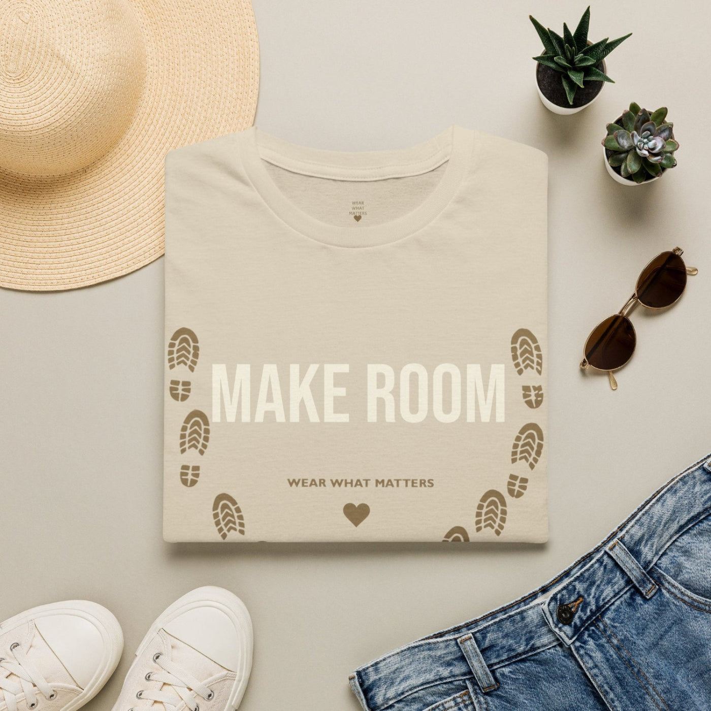 A folded cream-colored t-shirt with 'MAKE ROOM' and 'WEAR WHAT MATTERS' printed on the front, styled in a flat lay with a straw hat, white sneakers, blue jeans, sunglasses, and small potted succulents on a light gray background.