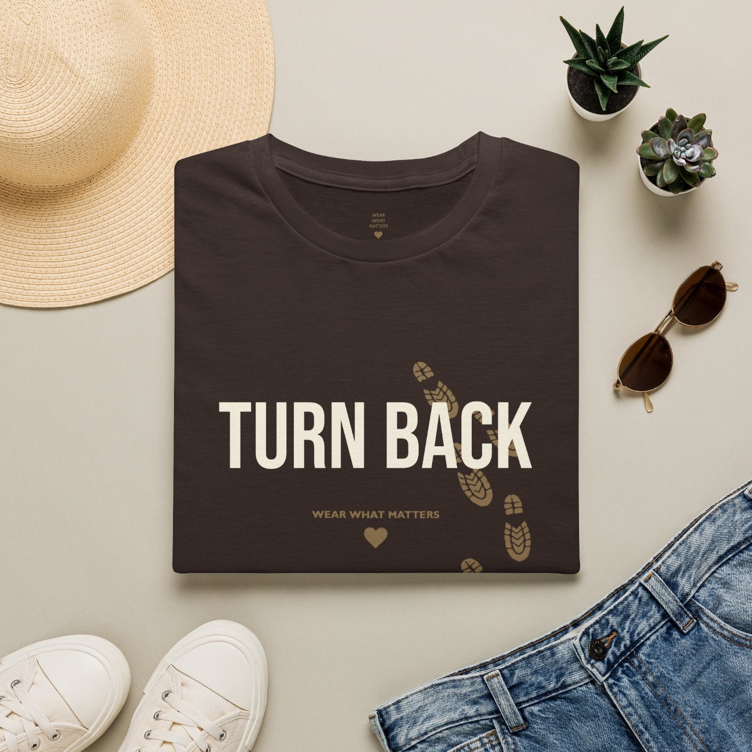 A folded black T-shirt is displayed in a styled flat lay with a graphic of four golf balls on tees. The shirt is surrounded by a straw hat, white sneakers, blue jeans, small potted succulents, and sunglasses on a light beige background.
Details