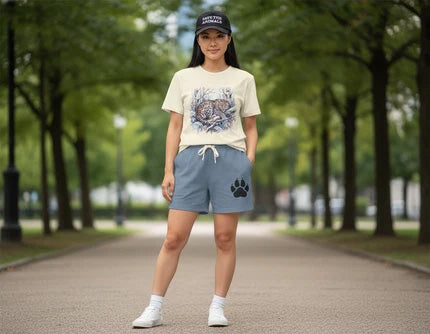A woman stands on a tree-lined path wearing a cream graphic t-shirt, slate blue drawstring shorts with a black paw print logo, white socks, and a black baseball cap.