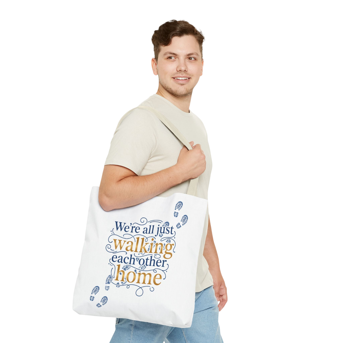 A man wearing a beige t-shirt and blue jeans carries a white canvas tote bag over his shoulder. The bag features the printed text 'We're all just walking each other home' in blue and orange lettering, decorated with blue footprints.