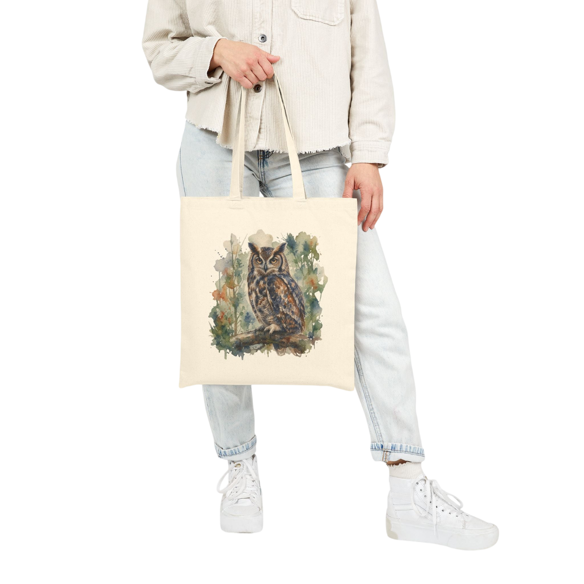 A person wearing a cream jacket, light-wash jeans, and white sneakers holds a cream canvas tote bag with a detailed owl illustration on the front, set against a plain white background.
