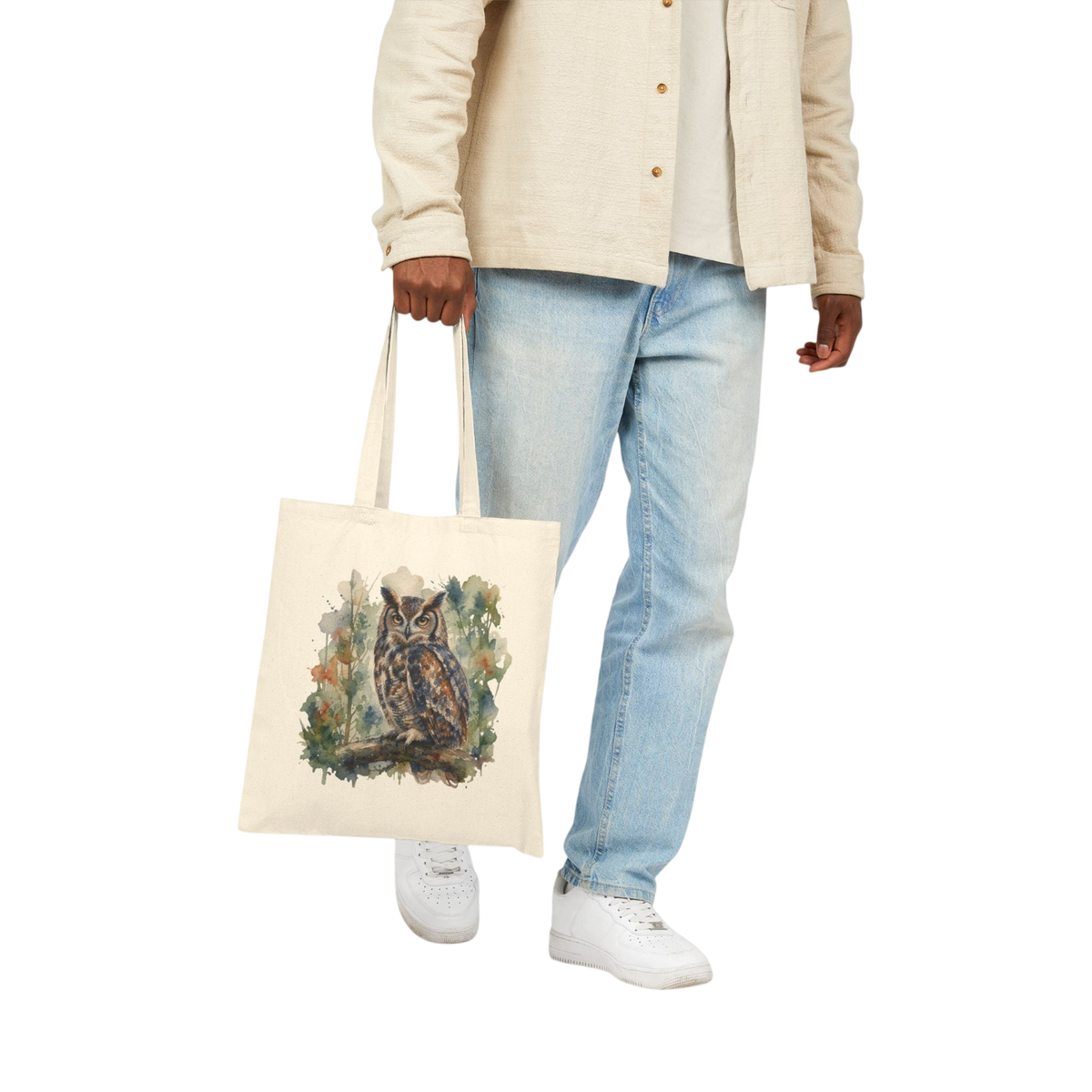 A person wearing a beige button-up shirt, light blue jeans, and white sneakers holds a cream canvas tote bag with a detailed owl illustration on the front, set against a plain white background.