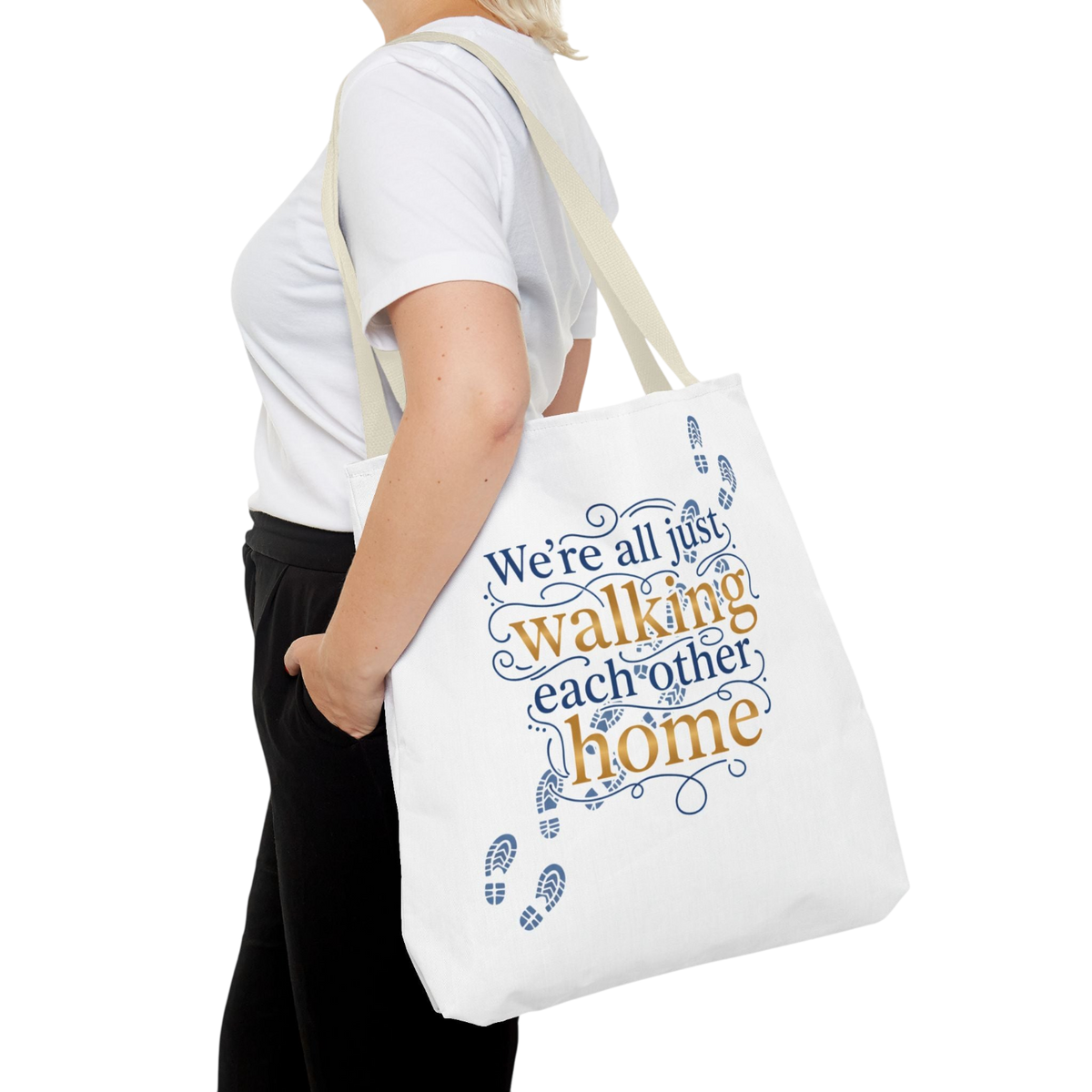 A person wearing a white t-shirt and black pants carries a white canvas tote bag with the printed phrase 'We're all just walking each other home' in blue and orange lettering, shown against a plain white background.