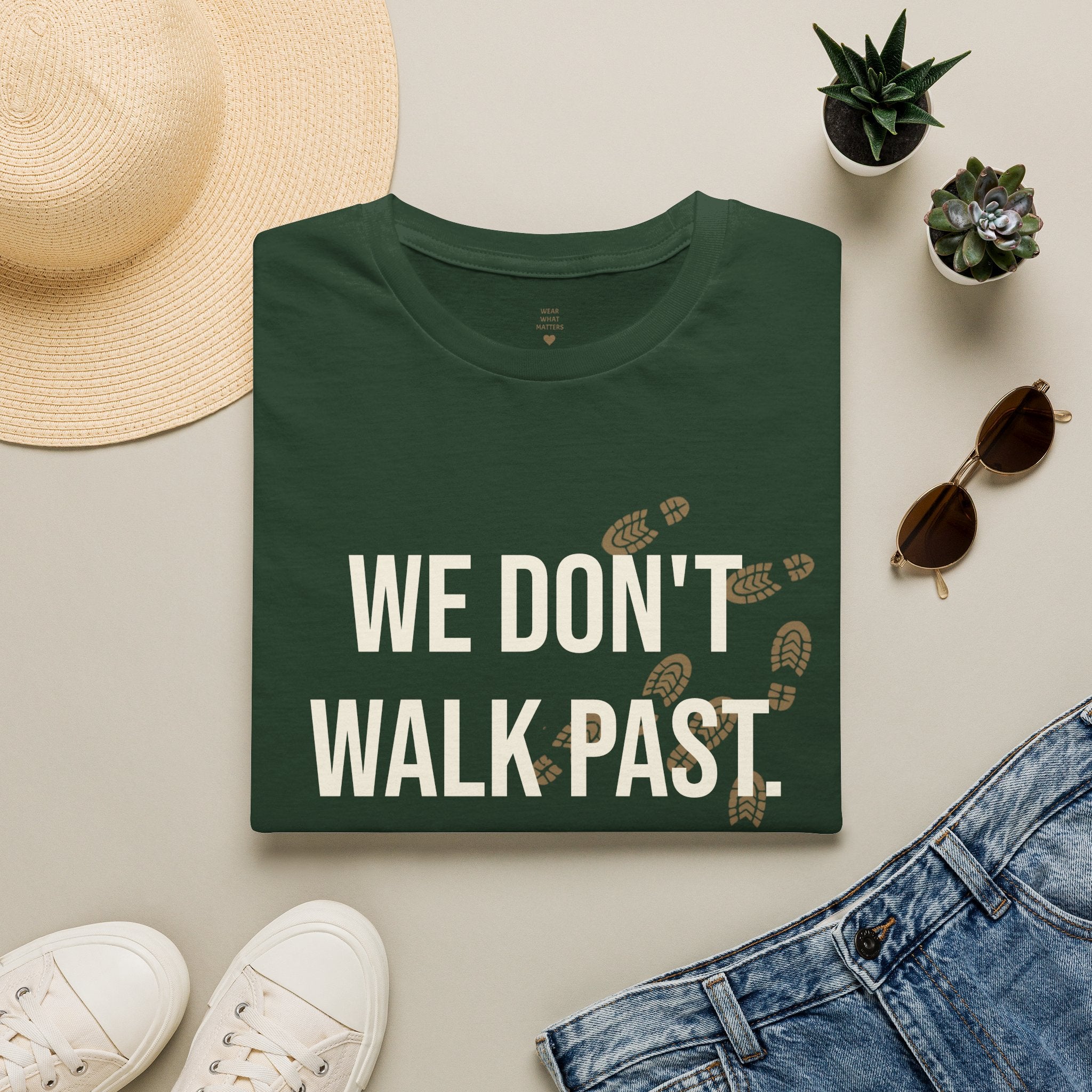A folded dark green T-shirt with the phrase 'WE DON'T WALK PAST.' in large white letters and small tan footprint graphics, styled in a flat lay with a straw hat, white sneakers, blue jeans, sunglasses, and two small potted succulents on a light beige background