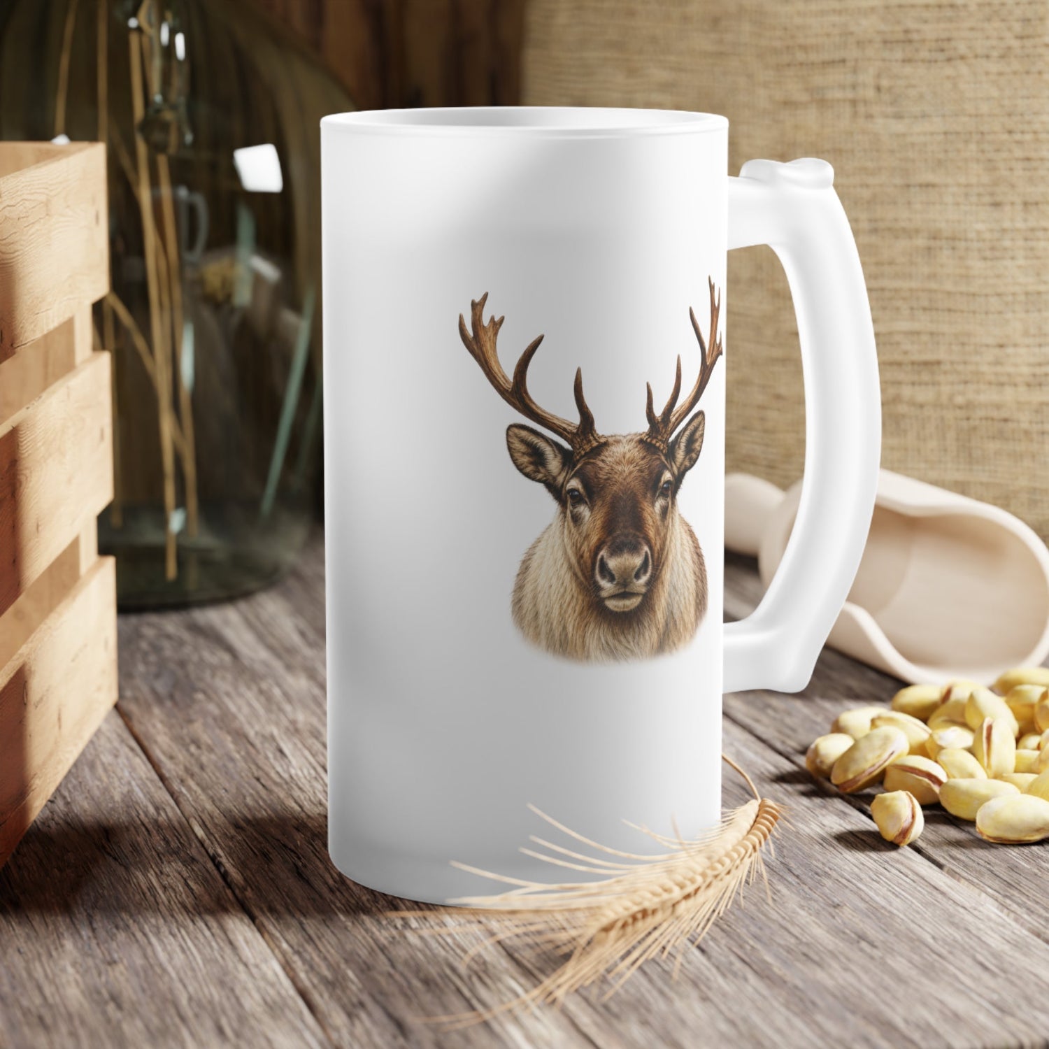 A frosted white ceramic mug with a detailed graphic of a reindeer head with large antlers, set on a rustic wooden table with a wheat stalk, pistachios, and a glass vase in the background.