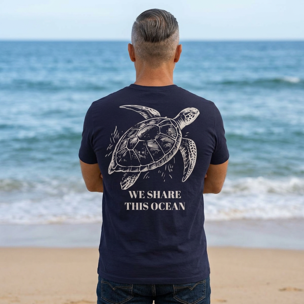 A man standing on a beach with his back to the camera, wearing a navy blue t-shirt with a white sea turtle graphic and the text 'WE SHARE THIS OCEAN' printed on the back.