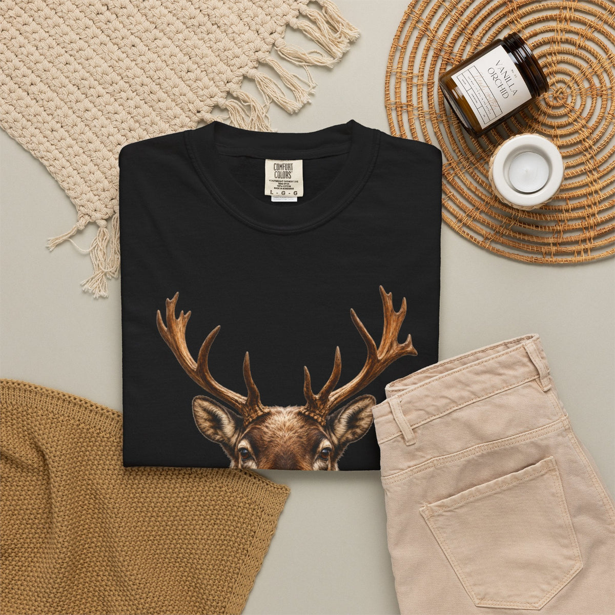 A folded black T-shirt with a detailed graphic of a deer head with large antlers, styled in a flat lay with beige pants, a knit blanket, a woven placemat, and a candle jar.