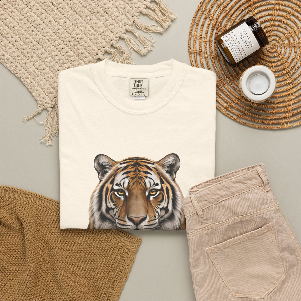A folded cream-colored t-shirt featuring a detailed graphic print of a tiger's face, styled in a flat lay with beige pants, a woven blanket, a candle jar, and a small cup on a neutral background.
Details