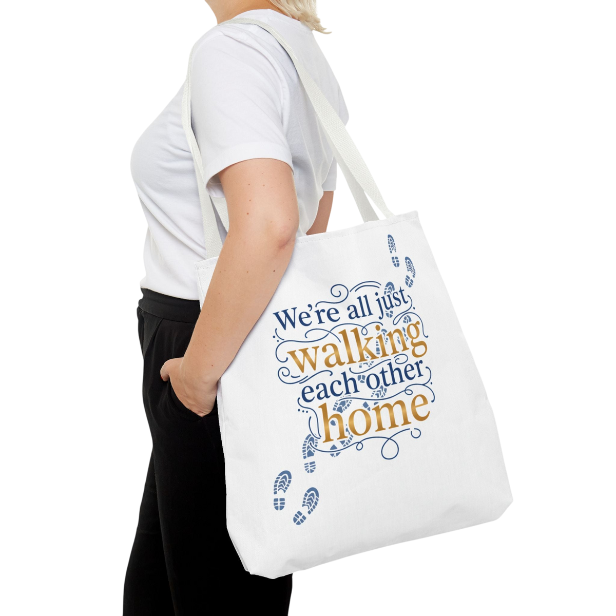 A woman wearing a white t-shirt and black pants carries a white canvas tote bag with the printed phrase 'We're all just walking each other home' in blue and orange lettering, shown against a plain white background.