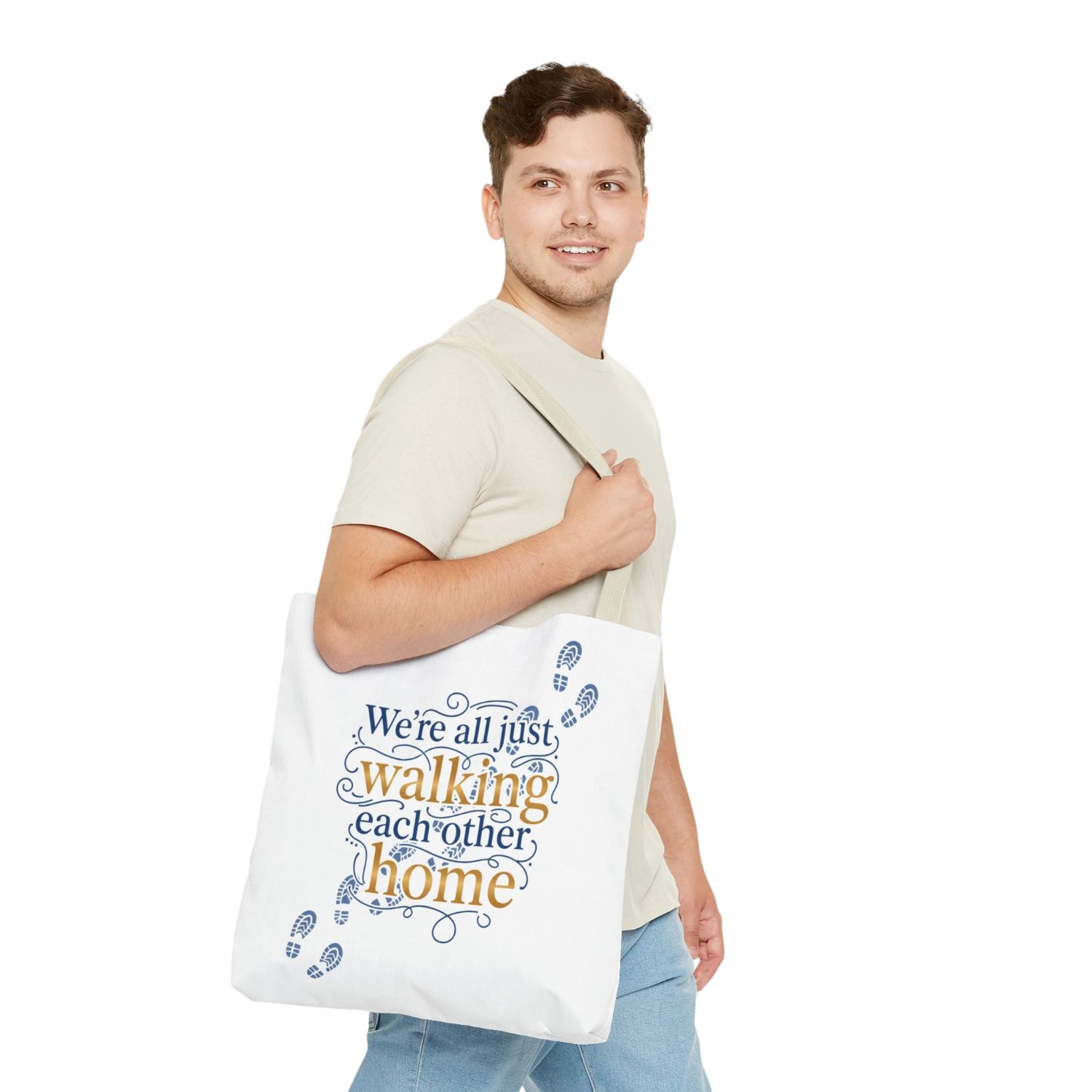 A man wearing a beige t-shirt and blue jeans carries a white canvas tote bag over his shoulder. The bag features the printed text 'We're all just walking each other home' in blue and orange lettering, decorated with blue footprints.
