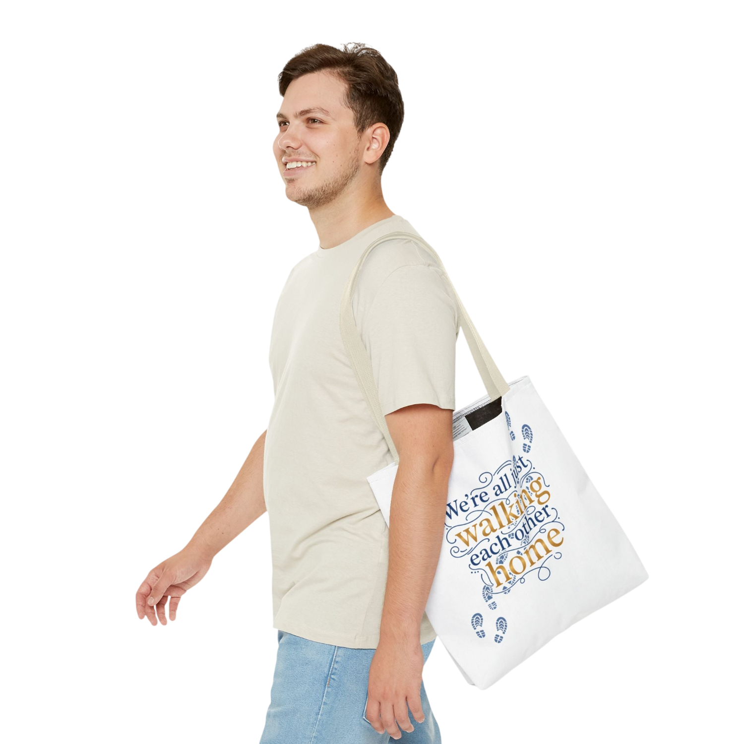 A man in a light beige t-shirt and blue jeans is shown from the side, carrying a white canvas tote bag over his shoulder. The tote bag features a blue and orange graphic with the text 'We're all walking each other home' and small blue footprints.