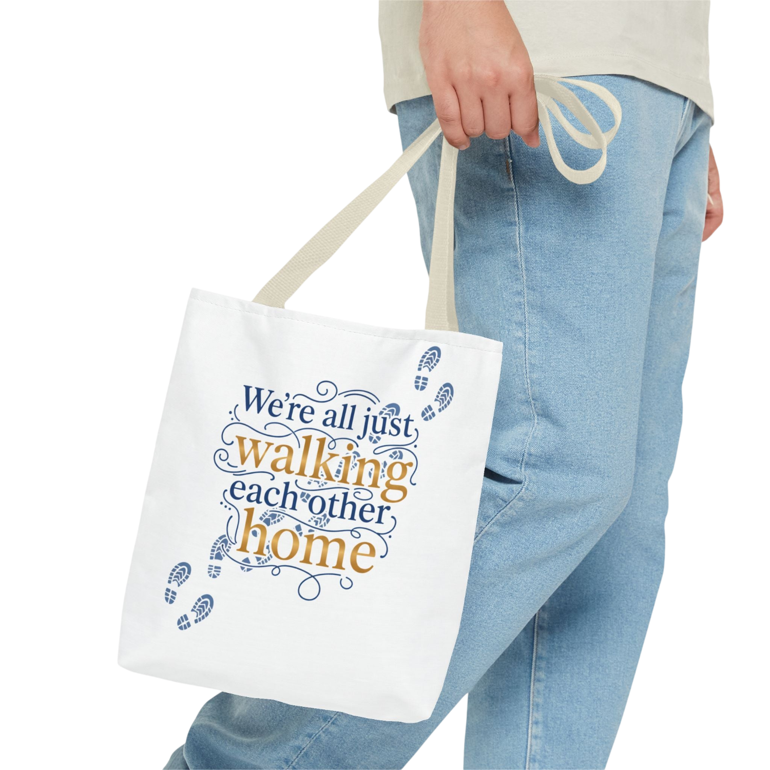 A white canvas tote bag with cream handles, featuring the printed phrase 'We're all just walking each other home' in blue and orange lettering with small blue footprints. The bag is held by a person wearing light-wash blue jeans against a plain white background.