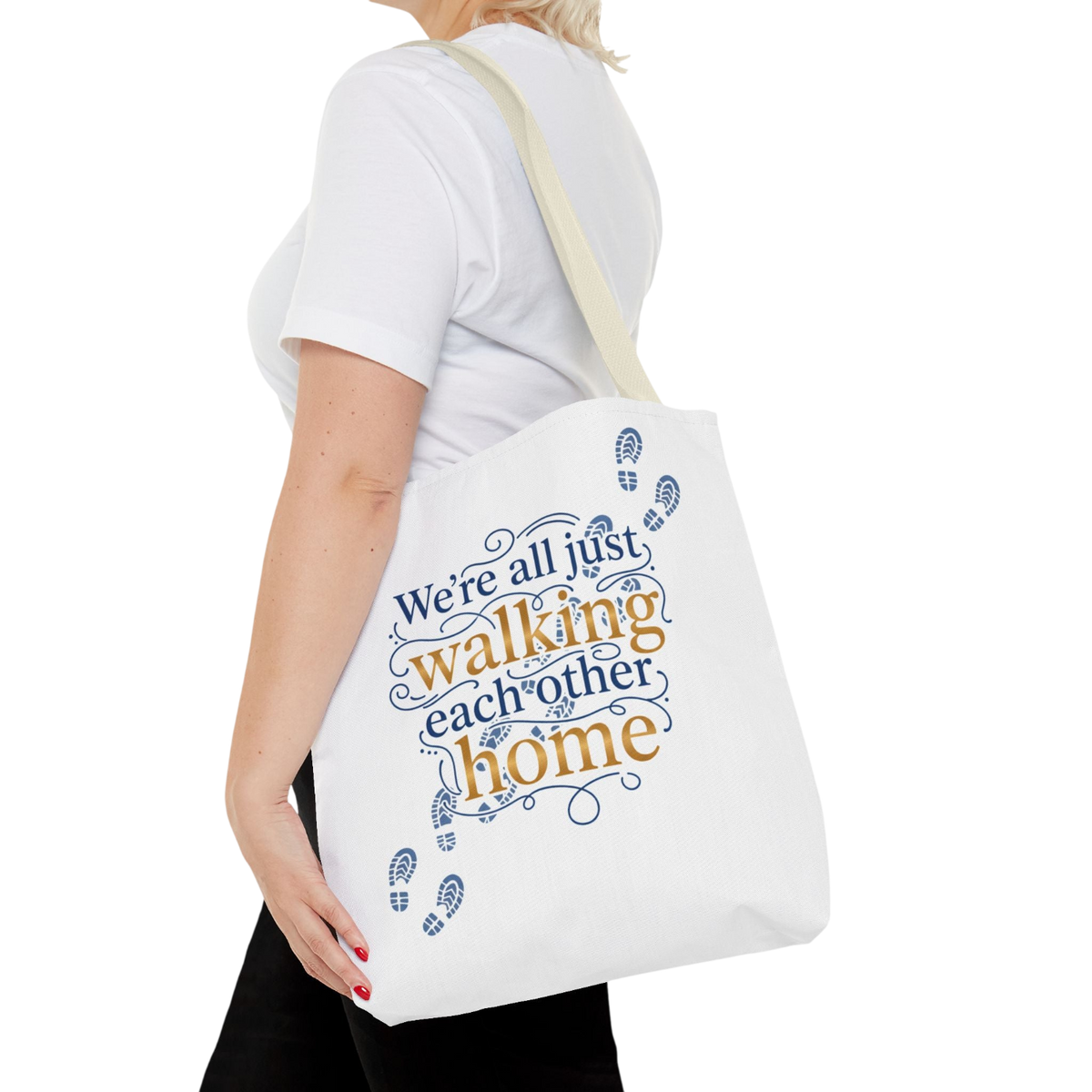 A woman wearing a white t-shirt and black pants carries a white canvas tote bag with a cream strap. The bag features a printed graphic with the text 'We're all just walking each other home' in blue and orange lettering, accented with blue footprint icons.