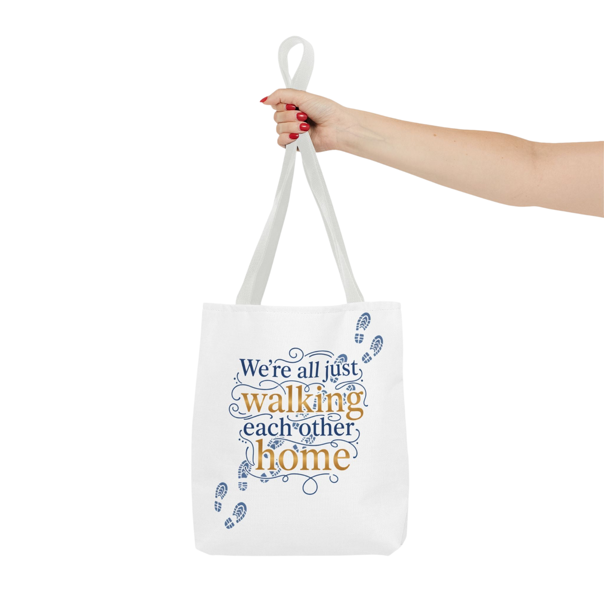 A white canvas tote bag with blue and orange text reading 'We're all just walking each other home' and small blue footprints, held by a person's hand against a plain white background.