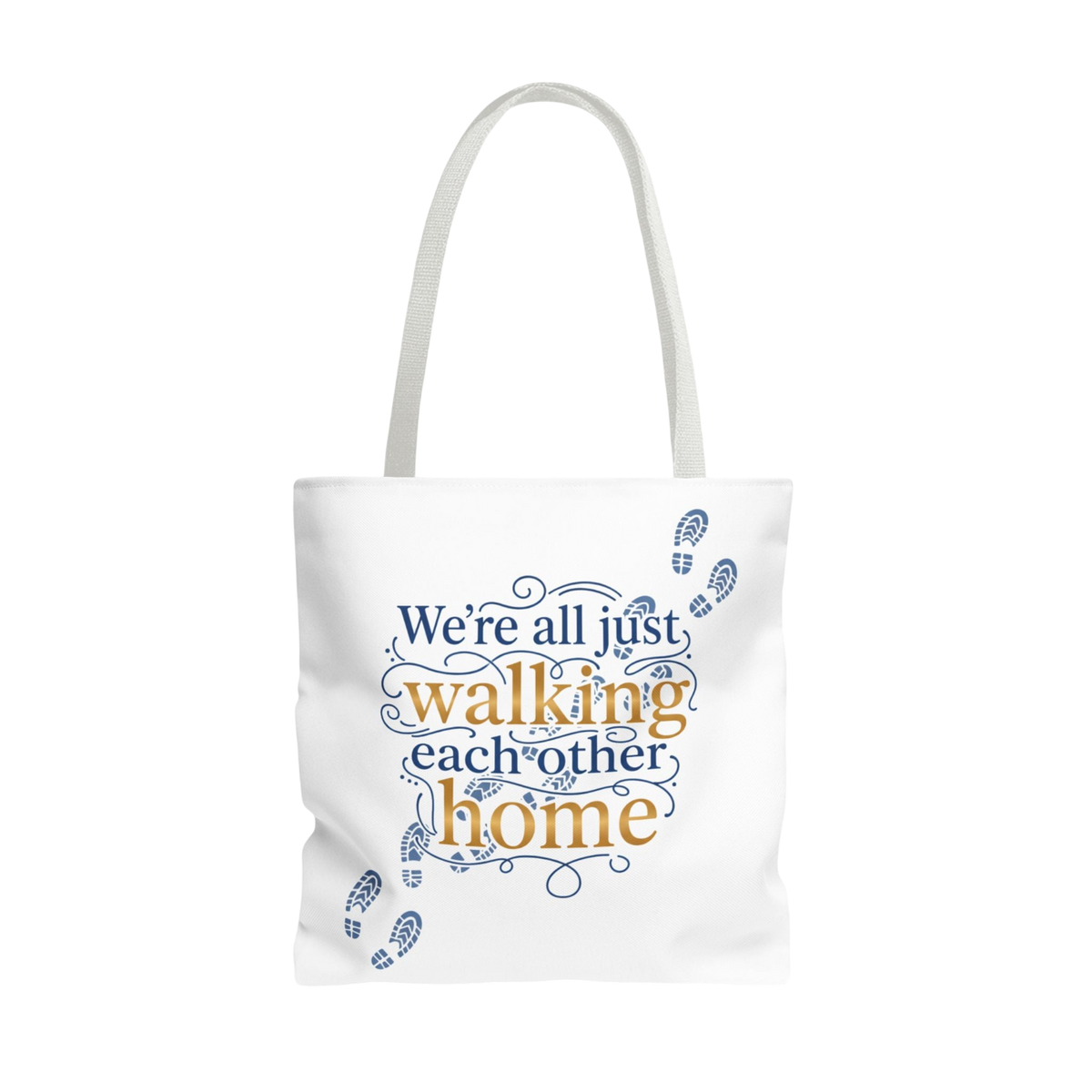 A white canvas tote bag with white shoulder straps, featuring a printed quote that reads 'We're all just walking each other home' in navy blue and mustard yellow lettering, accented with small blue footprint illustrations on a plain white background.