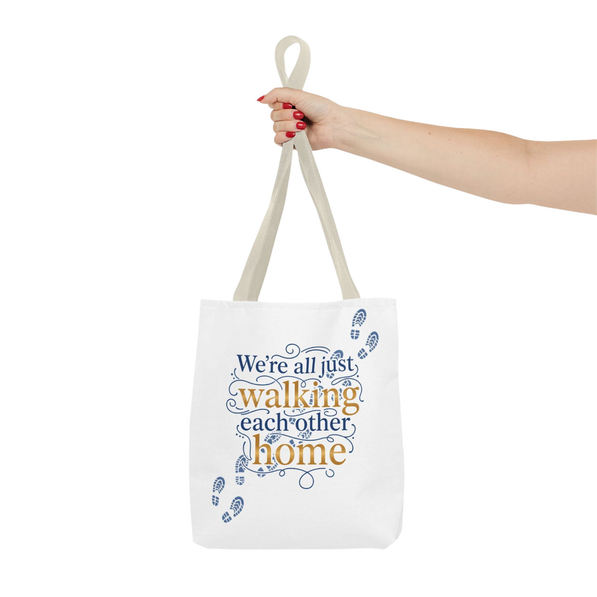A white canvas tote bag with beige handles, held by a person's hand against a plain white background. The bag features a printed design with the text 'We're all just walking each other home' in blue and orange lettering, decorated with blue footprints and swirls.